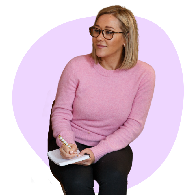 blond woman with glasses in pink sweater taking notes in a small notebook mid-conversation