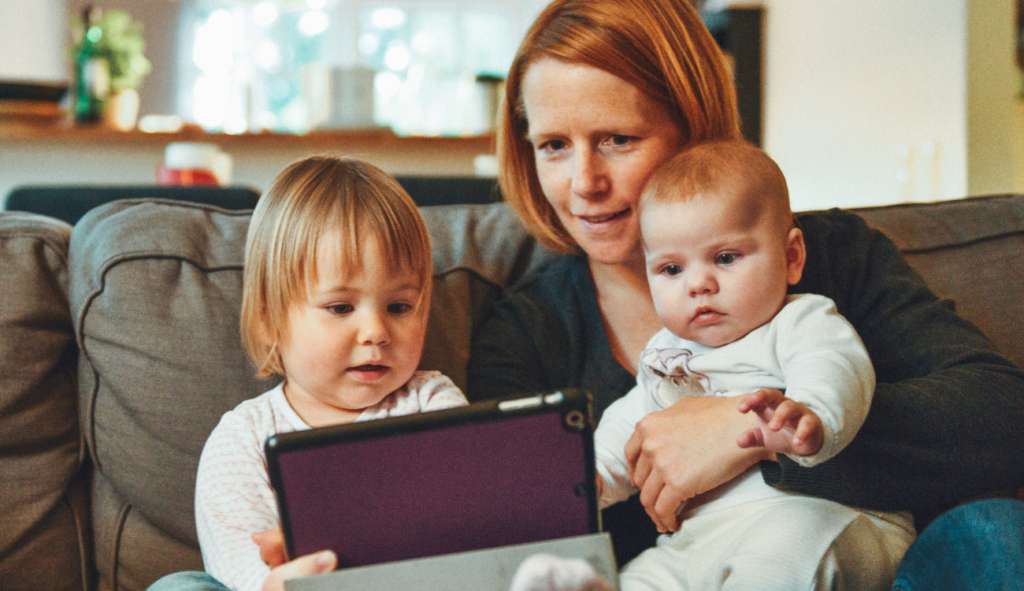 a mom sitting on the couch with two small children, showing the older one her tablet