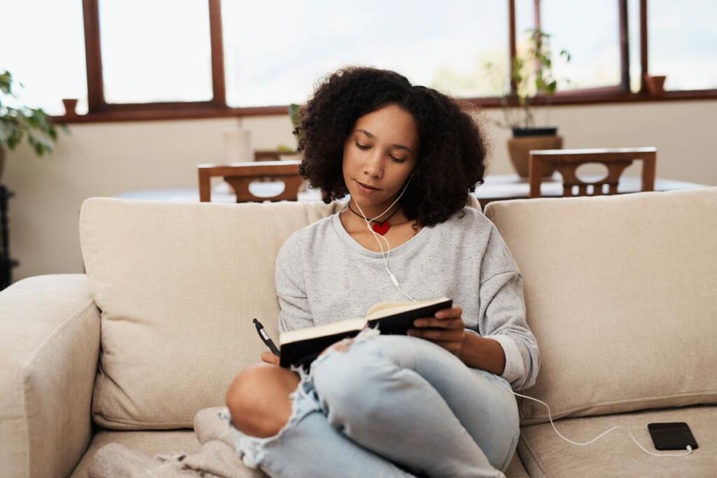 Woman listening to music with headphones and writing in journal on couch. 
