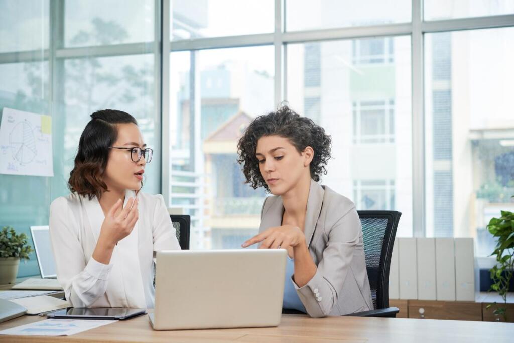 two women talking in an office, looking at the same computer together. good working relationships is important for short-term career goals.