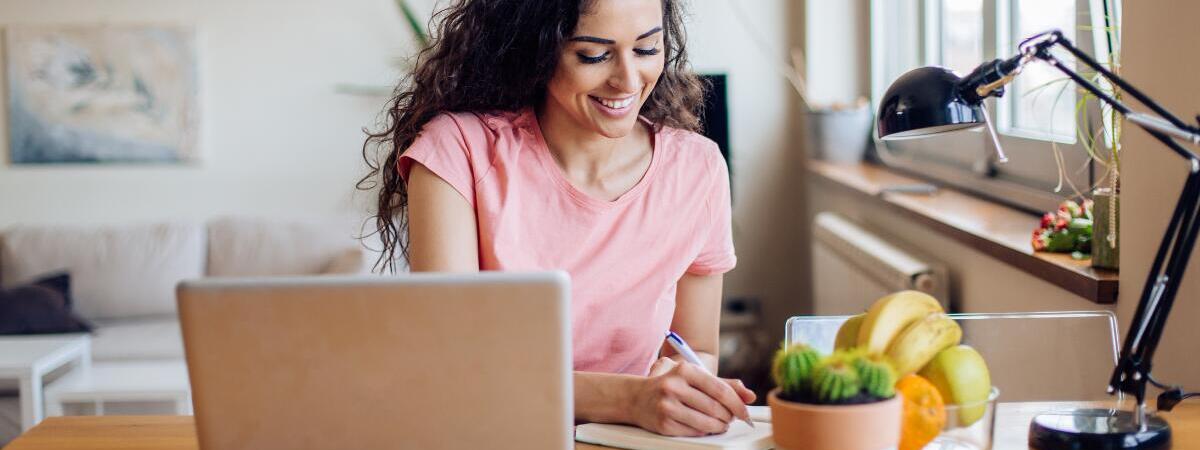 woman looking at computer while writing down notes