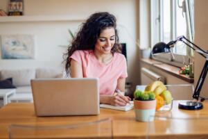 woman looking at computer while writing down notes