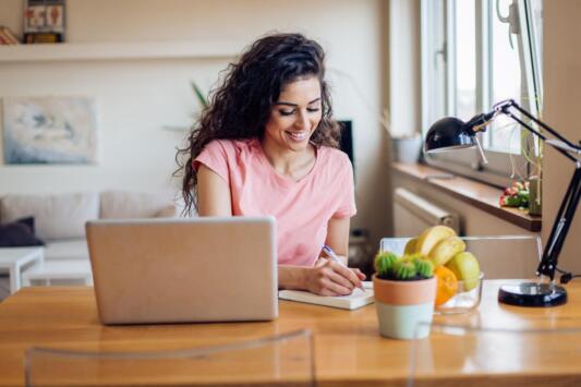 woman looking at computer while writing down notes